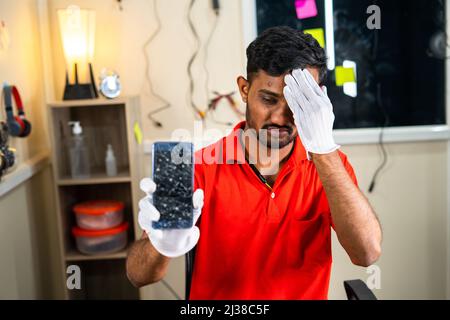 Besorgter Mechaniker, der in der Werkstatt ein kaputtes Mobiltelefon mit Händen auf der Stirn zeigt - Konzept der Fehler, des Facharbeiters und des beruflichen Berufs Stockfoto