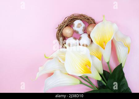 Bouquet von weißen gelben Callas mit Nest mit bemalten weißen und goldenen Eiern auf rosa Hintergrund. Frohe Ostern. Speicherplatz kopieren Stockfoto