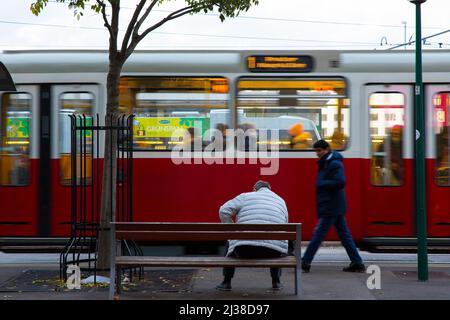 Straßenszene eines reifen Mannes, der auf einer Bank sitzt und auf die traditionellen öffentlichen Verkehrsmittel in Wien, die elektrische Straßenbahn, Österreich, wartet Stockfoto