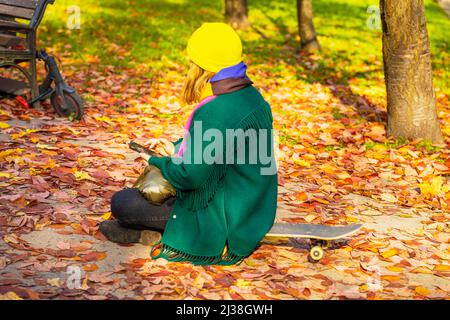 Junge unkenntliche Mädchen mit Telefon in Park mit Herbstblättern bedeckt, während sie auf dem Skateboard sitzen. Herbstsonniger Tag Stockfoto