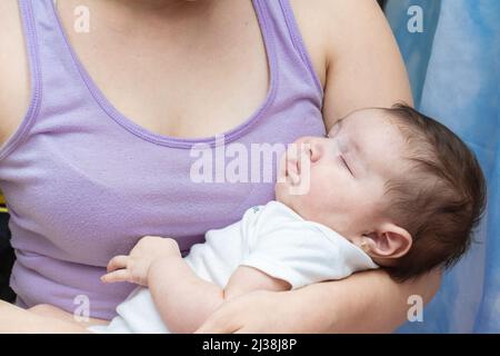 Schöne zwei Monate alte Latina Baby Mädchen schlafen auf den Armen ihrer Mutter. Nettes Baby Mädchen in einem weißen Jumpsuit gekleidet klammert an der Brust ihrer Mutter als s Stockfoto