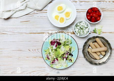 Frühstückstisch mit frischem Salat, hart gekochten Eiern, Cottage, Würstchen und Tomaten. Kopierraum, Holzhintergrund. Stockfoto