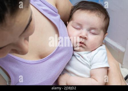 Schöne lateinische Baby-Mädchen schlafen zärtlich in den Armen ihrer Mutter, während Mutter weg schaut. Frau im Pyjama Pflege für zwei Monate altes Baby Mädchen. Konzept der Stockfoto