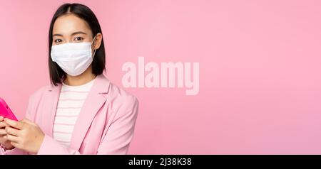 Asiatische Geschäftsfrau in medizinischen Gesichtsmaske mit Handy. Japanische Verkäuferin, Geschäftsfrau im Anzug, hält Smartphone und steht über Rosa Stockfoto