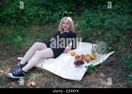 Hübsches blondes Mädchen auf einem Picknick in einem Wald oder Park mit Limonade, Früchten und Croissants. Sommerzeit, Ruhe, Entspannung. Millennial Generation. Stockfoto