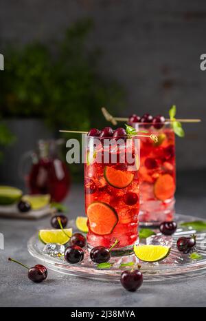 Kirschmojito. Sommercocktail in einem klaren Glas mit Kirschen, Limette, Pfefferminze und Eis auf grauem Hintergrund. Stockfoto