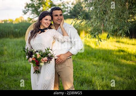 Niedliches Brautpaar umarmt und lächelt in einem grünen Park. Porträt der Braut und des Bräutigams in einem Spitzenkleid Stockfoto