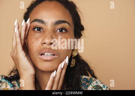 Junge schwarze Frau mit Ohrringen und Blick auf die Kamera isoliert auf beigefarbenem Hintergrund Stockfoto