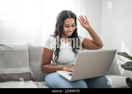 Attraktive junge afrikanische Frau, die am Laptop arbeitet, während sie zu Hause auf der Couch sitzt, sagt Hallo Stockfoto