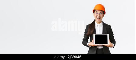 Aufgeregt und erstaunt lächelnd asiatische Bauingenieurin, Architekt in Sicherheitshelm und Business-Anzug, zeigt digitale Tablet-Bildschirm mit erstaunt Stockfoto