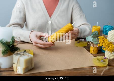 Beschnittene Ansicht der Kunsthandwerkerin, die handgemachte Kerzen neben Seife und Bastelpapier auf dem Tisch hält Stockfoto