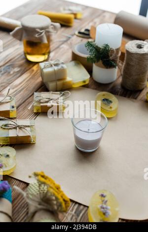 Handgemachte Kerze aus Glas in der Nähe von Seifenstücken und Bastelpapier auf Holzoberfläche Stockfoto