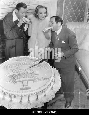 Dietrich Fetes Lubitsch -- sie beim Kuchenschneiden. Die Torte war mit 25 Kerzen geschmückt und trug die Inschrift „Herzlichen Glückwunsch Ernst, Marlene“. Die gesamte Filmgesellschaft von mehr als 50 Personen schloss sich der Partei an. Vor genau 25 Jahren begann Ernst Lubitsch 1912, sein erstes Bild im Berliner Stadtteil Tempelhof zu inszenieren. Zu Ehren des Anlasses gab Marlene Dietrich eine Party für ihn – am Set, wo er sie und Herbert Marshall in Paramount's 'Angel' dirigiert. 15. November 1937. (Foto von C.E. Gatward) Stockfoto