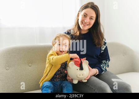 Fröhliche Mutter Teaching Little Baby wie man Geld sparen Halten Sparschwein sitzen auf Sofa zu Hause Stockfoto