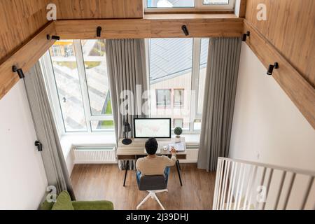 Rückansicht des cleveren und konzentrierten kaukasischen Mannes, der Papierkram macht. Ein Mann schaute durch Papier und saß am Schreibtisch mit einem Computer im Büro. Man prüft das Berichtskonzept Stockfoto