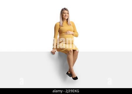In voller Länge Porträt einer Schwangeren in einem gelben Kleid sitzt auf einem leeren Panel isoliert auf weißem Hintergrund Stockfoto