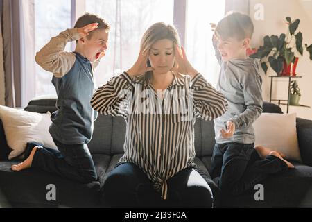 Unglückliche kaukasische Mutter leiden unter Kopfschmerzen, die müde vom Spielen eines frechen kleinen Sohnes gestresst werden. Stockfoto