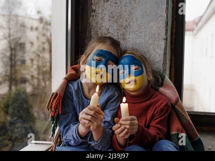 Zwei kleine Mädchen mit ukrainischen Flaggen auf ihren Gesichtern beten für die Ukraine und halten brennende Kerzen in den Händen, eine Geste des Glaubens und h Stockfoto