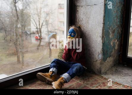 Ein kleines ukrainisches Patriotin mit der Flagge der Ukraine im Gesicht betet am Fenster. Kinder sind gegen den Krieg in der Ukraine. Stockfoto