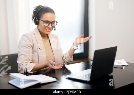 Frau mittleren Alters, die ein schnurloses Headset trägt und einen Laptop verwendet, um mit Kunden oder Kollegen zu kommunizieren, die am Schreibtisch im modernen Büro sitzen. Weibliche Unterstützungsarbeiterin spricht online Stockfoto