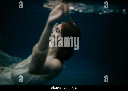 Frau schwimmt im Wasser mit geschlossenen Augen und trägt ein glitzerndes Kleid Stockfoto