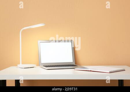 Leuchtende Lampe, moderner Laptop und geöffnetes Notebook auf dem Tisch neben der Farbwand Stockfoto