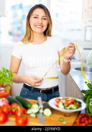 Die junge Latino-Frau misst ihre Taille mit einem Maßband in der Küche Stockfoto