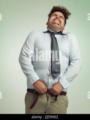 Bereit, seine Hose zu schließen. Humorvolle Studioaufnahme eines übergewichtigen Geschäftsmannes, der versucht, seine Hose zu knöpfen. Stockfoto