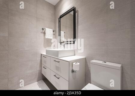 Badezimmer mit weißem Porzellanwaschbecken, quadratischem Spiegel mit schwarzem Metallrahmen, glänzend weißem Hängeschrank und Chromarmaturen Stockfoto