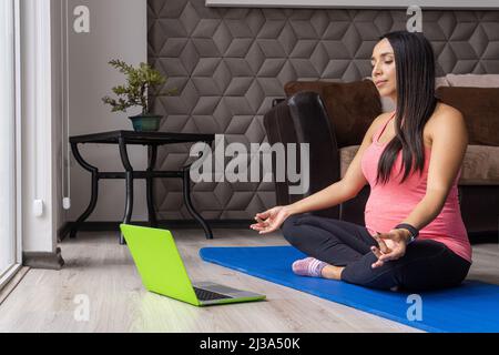 Junge, Schwangerische Lateinerin, die online Yoga- und Meditationskurse auf einem Laptop erhält Stockfoto