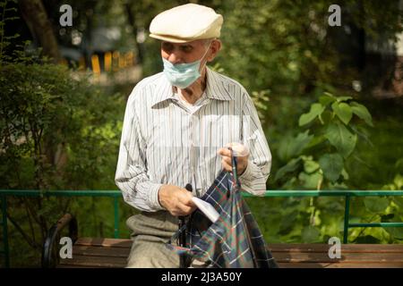 Der alte Mann sitzt im Sommer auf der Bank. Rentner in Russland in der Nähe des Hauses. Mann mit medizinischer Maske. Opa in Hemd und Mütze. Stockfoto