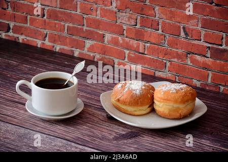 Zwei Donuts mit Puderzucker auf einem Teller und eine Tasse schwarzen Kaffee auf einem Holztisch gegen eine Ziegelwand. Nahaufnahme. Stockfoto