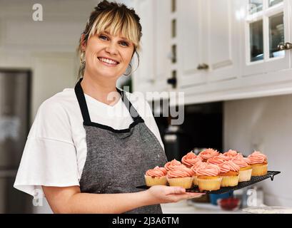 Backen Sie sich glücklich. Ein kurzer Schuss einer Frau, die zu Hause in ihrer Küche frisch gebackene Cupcakes hält. Stockfoto