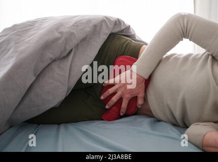 Berlin, Deutschland. 30. März 2022. ABBILDUNG - Eine Frau hält eine Wärmflasche an ihren Unterbauch, während sie im Bett liegt. (Posed scene) viele Betroffene der Bauchkrankheit Endometriose leiden im Alltag unter schweren Symptomen und damit verbundenen Einschränkungen. (To dpa: 'Mysterium Endometriose - Wenn die Zeit zur Qual wird') Credit: Annette Riedl/dpa/Alamy Live News Stockfoto