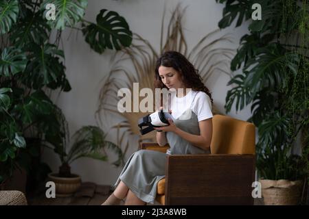 Neugierige junge Frau, die modernes VR-Headset untersucht, während sie im gemütlichen Hausgarten sitzt Stockfoto