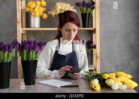 Eine junge Floristin in einer blauen Schürze überprüft Bestellungen auf einem Mobiltelefon, während sie in einem Geschäft mit frischen Blumen an einem Tisch in Glasvasen sitzt. Stockfoto