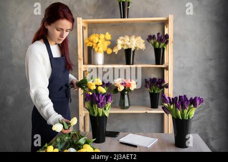 Ein kleines Geschäft. Eine Frau Floristin in einem Blumenladen in der Nähe einer Vitrine mit Tulpen in einer Schürze sammelt einen Strauß von Tulpenblumen. Blumenlieferung Stockfoto