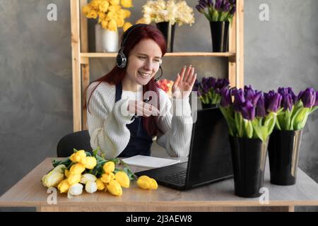 Eine Callcenter-Agentin mit Headset arbeitet an einer Hotline hinter einem Laptop. Weibliche Floristin im Blumenladen in der Nähe von Vitrine mit Tulpen im Vorfeld, die bestellen Stockfoto