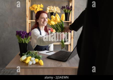 Ein junger Mann kauft für den Urlaub eines Mädchens in einem gemütlichen Blumenladen einen schönen Blumenstrauß aus Tulpenblumen. Floristik und Herstellung von Blumensträußen in einem Blumenladen Stockfoto