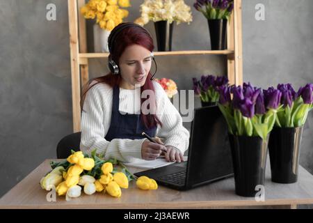 Eine Callcenter-Agentin mit Headset arbeitet an einer Hotline hinter einem Laptop. Weibliche Floristin im Blumenladen in der Nähe von Vitrine mit Tulpen im Vorfeld, die bestellen Stockfoto