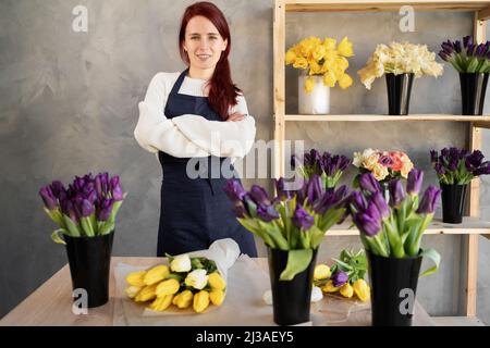 Das junge kaukasische Verkaufsmädchen mit gekreuzten Armen in einer Arbeitsschürze lächelt fröhlich, während es in einem Blumenladen an einem Schaufenster steht. Stockfoto