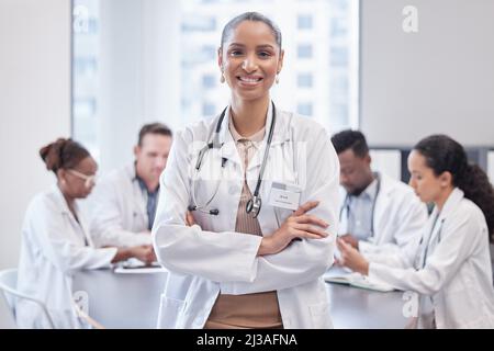 Selbstbewusst mit einem tollen Team hinter mir. Beschnittenes Porträt einer attraktiven jungen Ärztin, die mit gefalteten Armen im Sitzungssaal steht Stockfoto