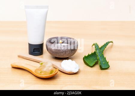 Eine Nahaufnahme von Cremetöhrchen, grünen Aloe-Vera-Schalen, Aloe-Vera-Gelee und weißer Creme in Holzlöffeln auf einem Holztisch mit verschwommenem Hintergrund Stockfoto