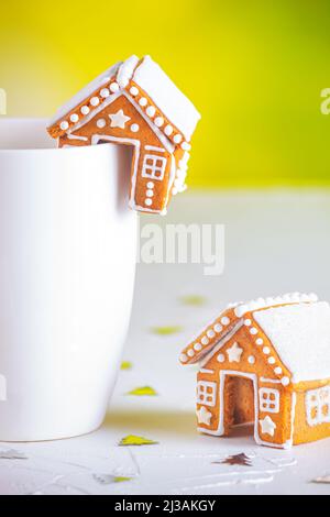 Lebkuchenhütten und eine Tasse Milch auf einer weißen Betonoberfläche mit Funkeln, Nahaufnahme. Weihnachtsfeier Konzept Stockfoto