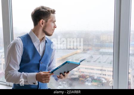 Der junge bärtige Geschäftsmann arbeitet im Büro, notiert sich im Notizbuch, analysiert Informationen, macht Geschäftsplan, während er am Fenster auf der Fensterbank sitzt. Freiberuflicher Unternehmer, der von zu Hause aus arbeitet. Stockfoto