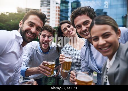Wir feiern das Jahresende. Porträt einer fröhlichen Gruppe junger Arbeitskollegen, die sich beim Biertrinken draußen für ein Foto nahe kommen Stockfoto