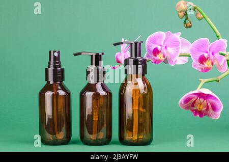 Braune Kosmetikflaschen mit Spender auf grünem Hintergrund mit Orchideenblumen Stockfoto