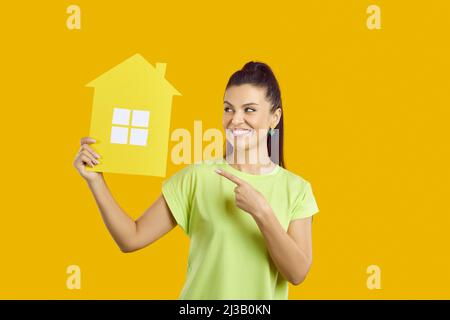 Positiv lächelnde Frau mit braunen Haaren, die mit dem Finger auf das Papierspielzeughaus in der Hand zeigt, Mietung und Verkauf von Wohnungen, Immobilienagentur. Stockfoto