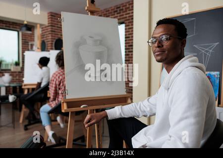 Porträt eines lächelnden afroamerikanischen Studenten, der eine Kunstklasse bescheinigte, die neue künstlerische Fähigkeiten für persönliches Wachstum entwickelte. Diverse Team Zeichnung Vase Modell arbeiten an Grafik-Technik. Kreatives Konzept Stockfoto