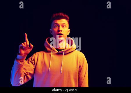 Junger, hübscher Mann mit Kapuzenpullover, isoliert über dunklem Studiohintergrund in Neonlicht. Konzept der Emotionen, Mode Stockfoto